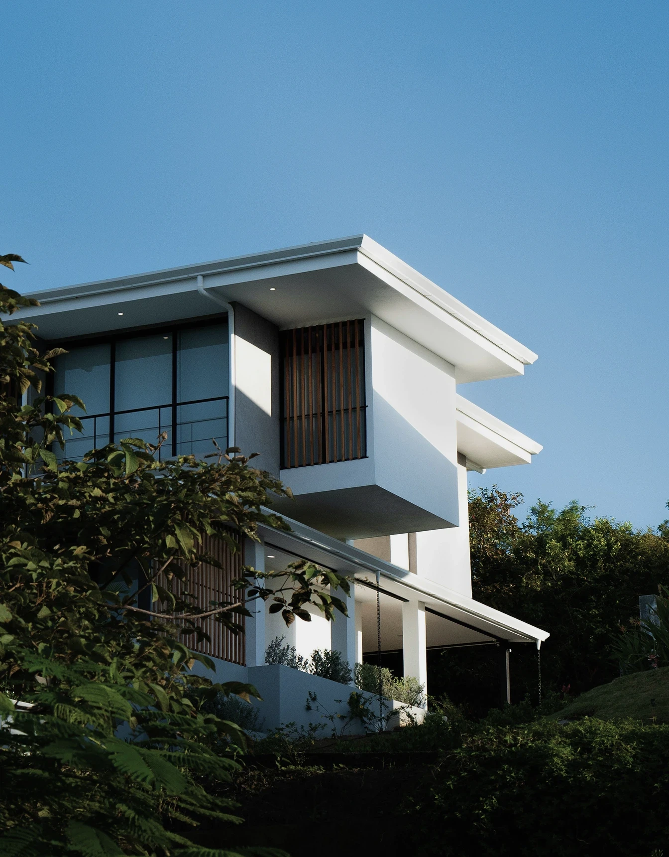 Modern white house with large overhanging roof, wooden slats, and expansive glass windows. Surrounded by lush greenery under a clear blue sky.