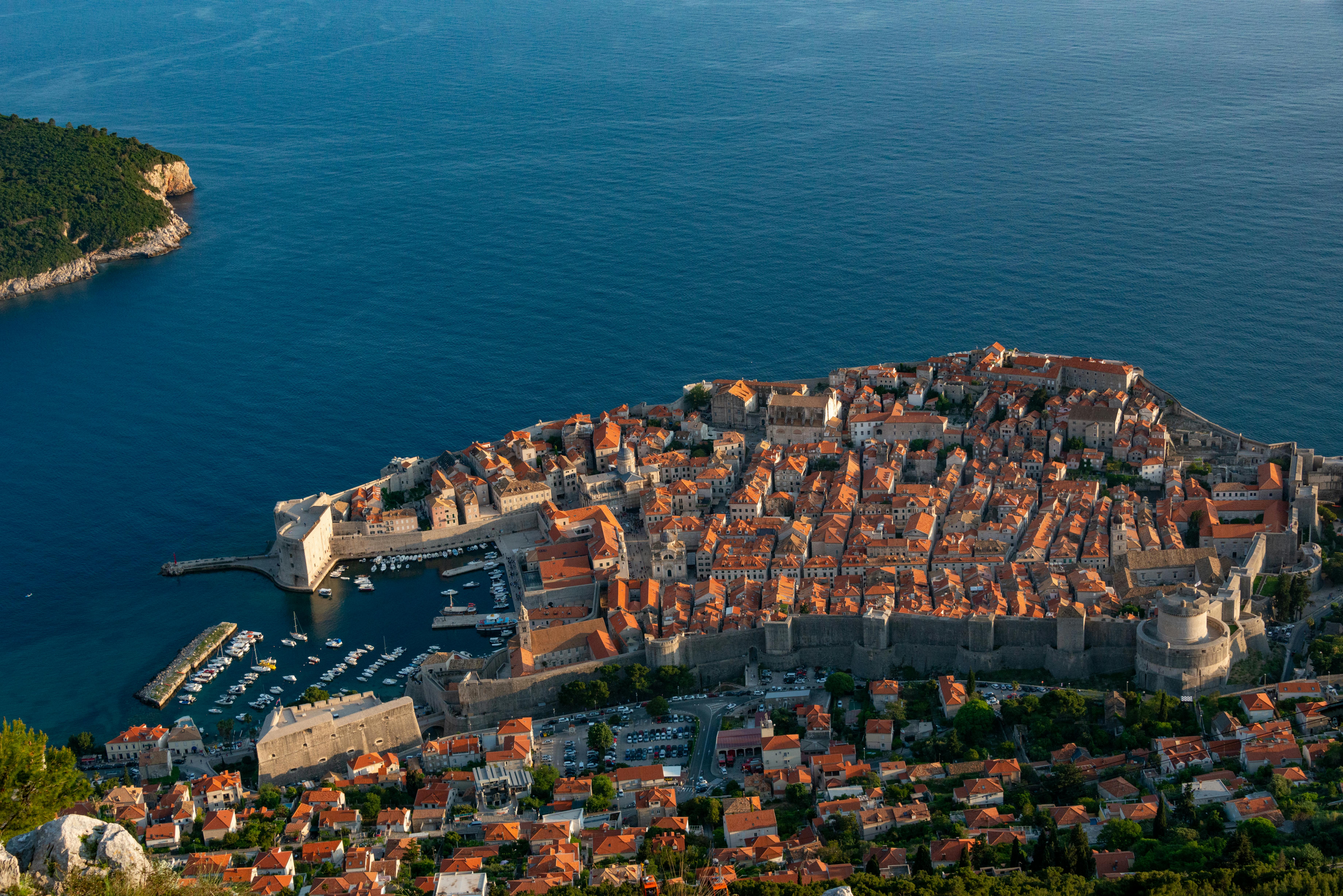 Aerial view of a coastal city with a harbor, surrounded by blue water and green hills in the background.