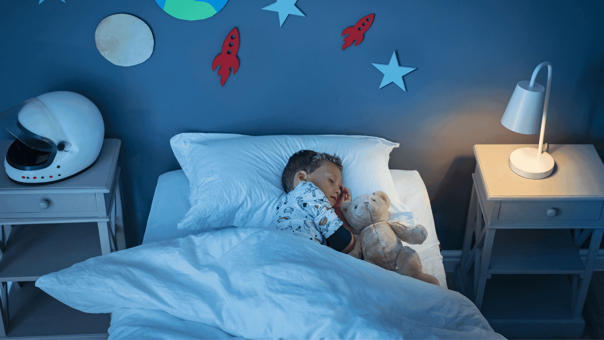 A boy asleep in bed with a night light on a bedside stool and a helmet on the other side, with space artwork on the walls.