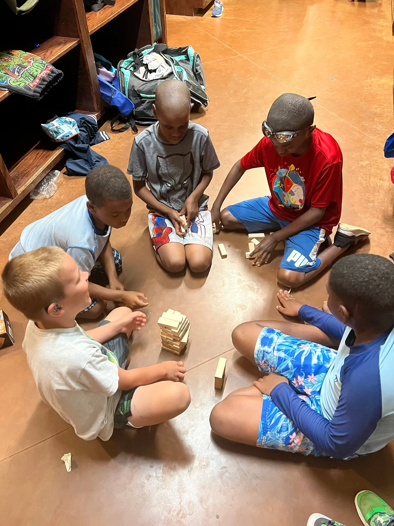 Five boys sit in a circle on the floor playing a game with wooden blocks.