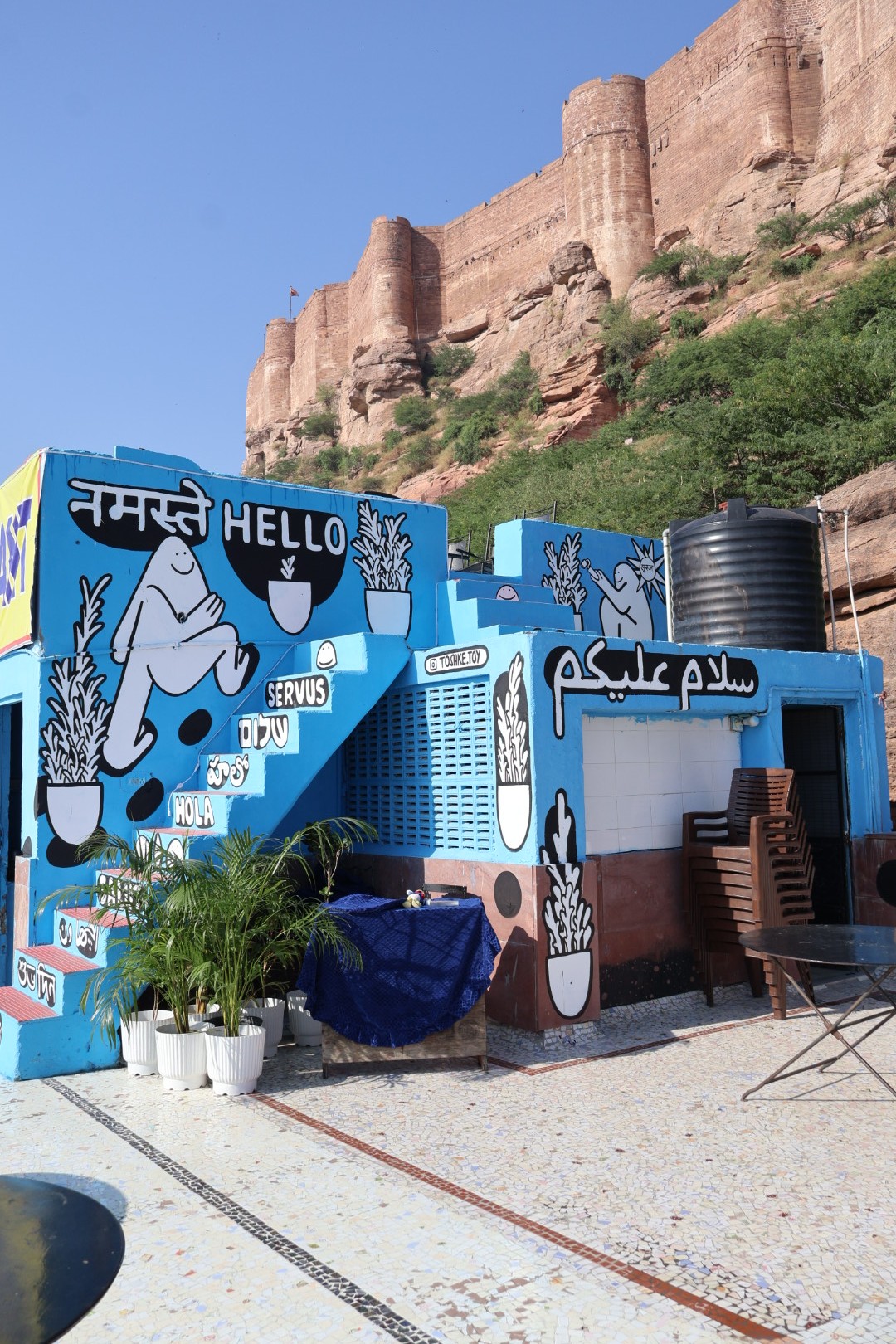 Mural painting with joyful cartoon characters and the word ‘Hello, नमस्ते, سلام عليكم’ painted on a terrace of a blue building near Jodhpur Fort.