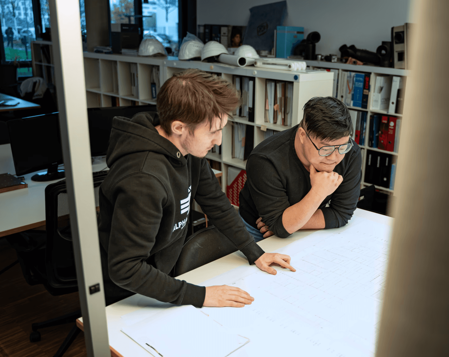 Zwei Mitarbeitende der kba Architekten und Ingenieure GmbH beugen sich über großformatige technische Baupläne und analysieren Details; im Hintergrund stehen Regale mit Ordnern und Sicherheitshelmen in einem modernen Büro.