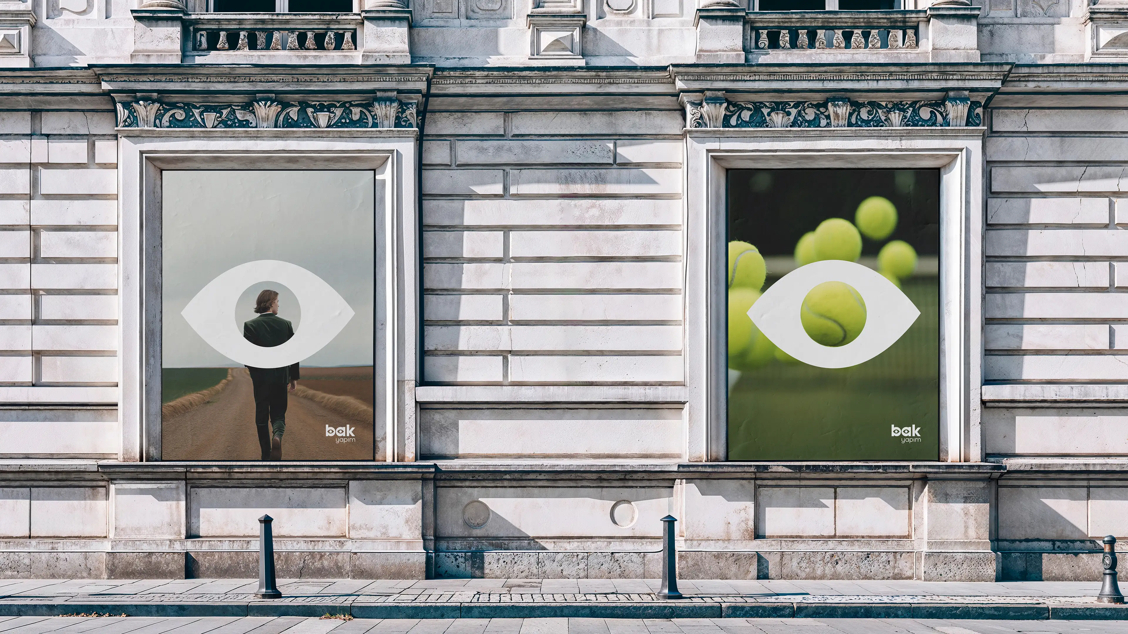 Exterior view of a classical stone building featuring large window displays with the Bak Yapım eye symbol and minimalist brand posters.