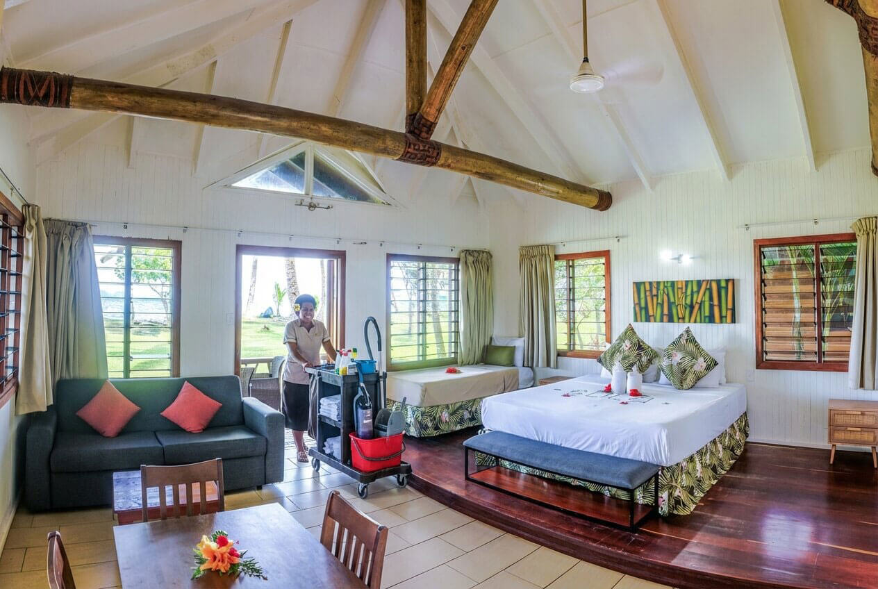 A smiling staff member cleans a spacious beachfront bure at Uprising Beach Resort, featuring a bed, sofa, and ocean view.