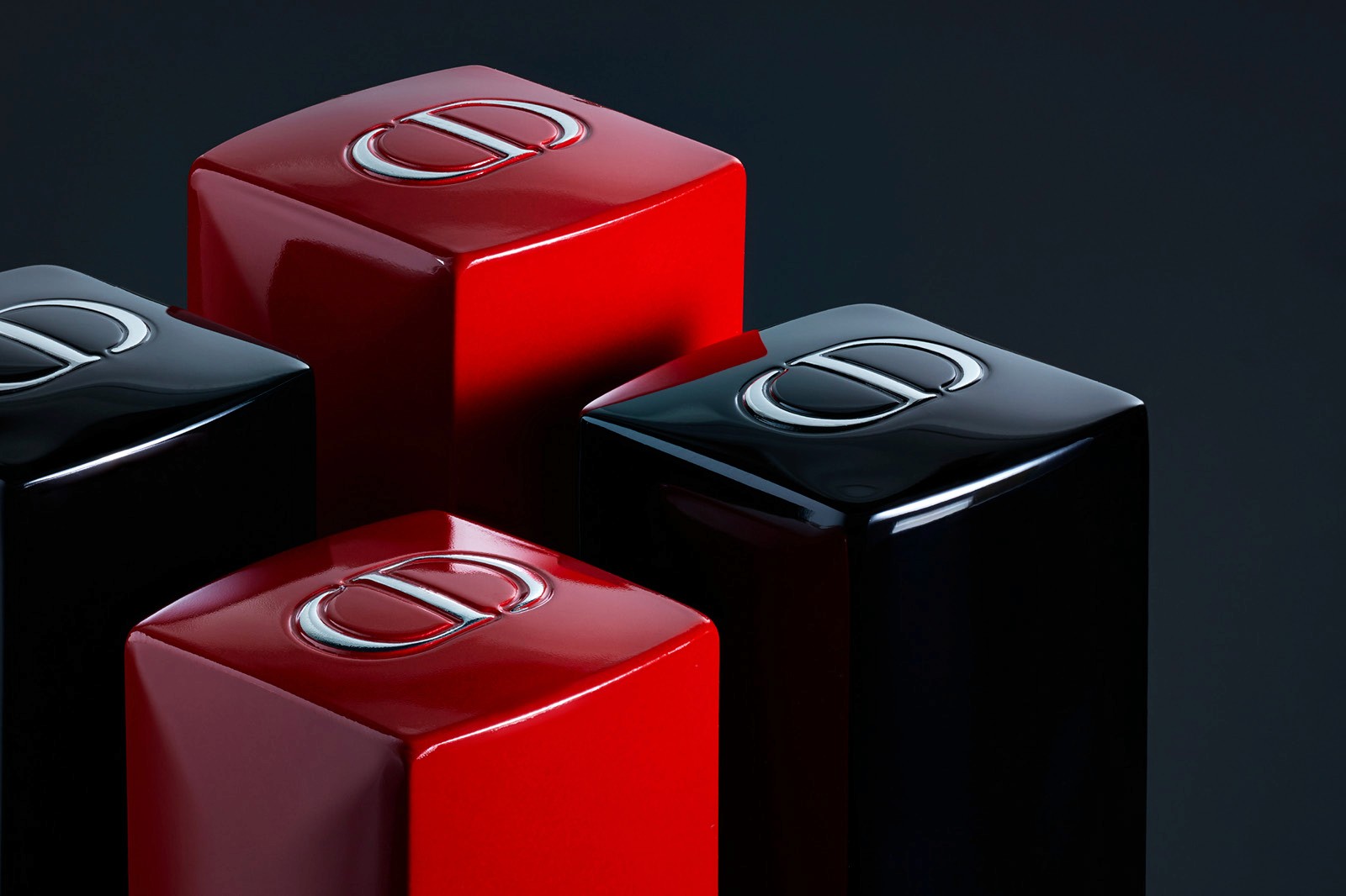 Three glossy make-up products, two red and one black, are arranged creatively against a dark background with the Christian Dior monogram on the top.