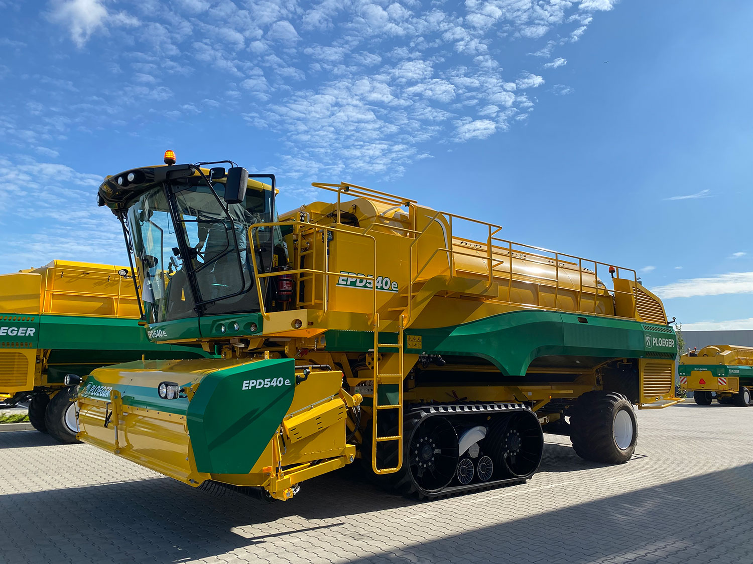 Side view of a Ploeger harvester. It is bright yellow with green accents. The sky is blue with dappled cloud.