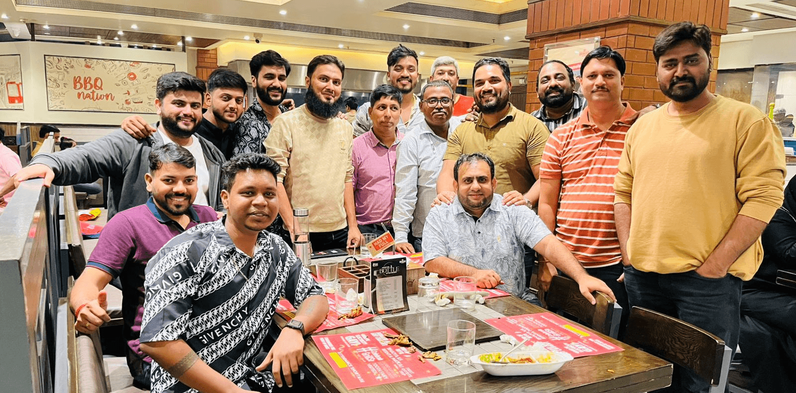 A group of people smiling together around a table, enjoying food in a lively indoor setting.