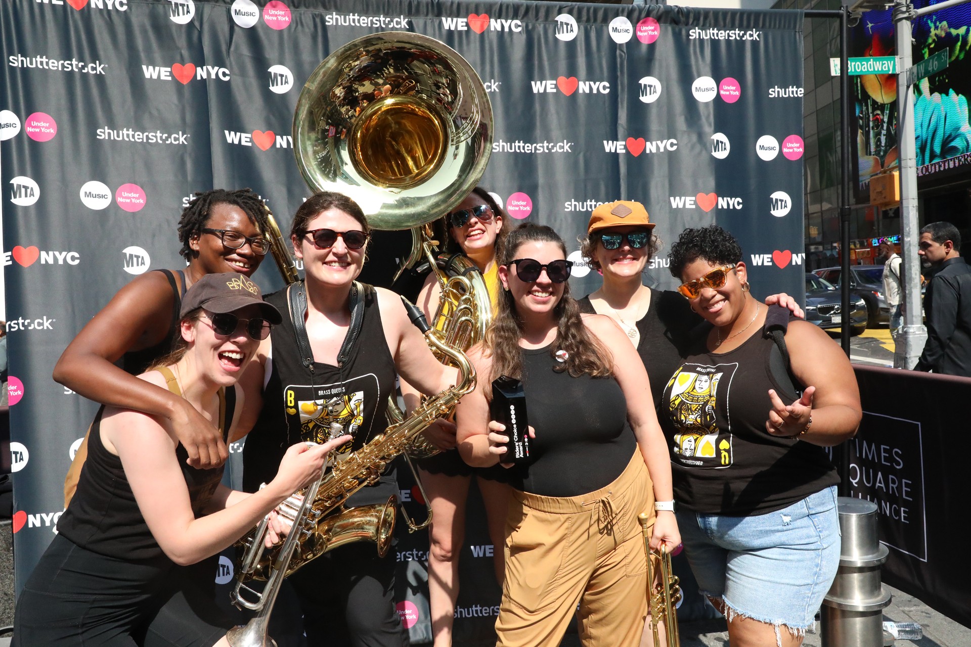 6 members of Brass Queens stand in a group with musical instruments in front of a wall of logos for the MTA and We Love NYC