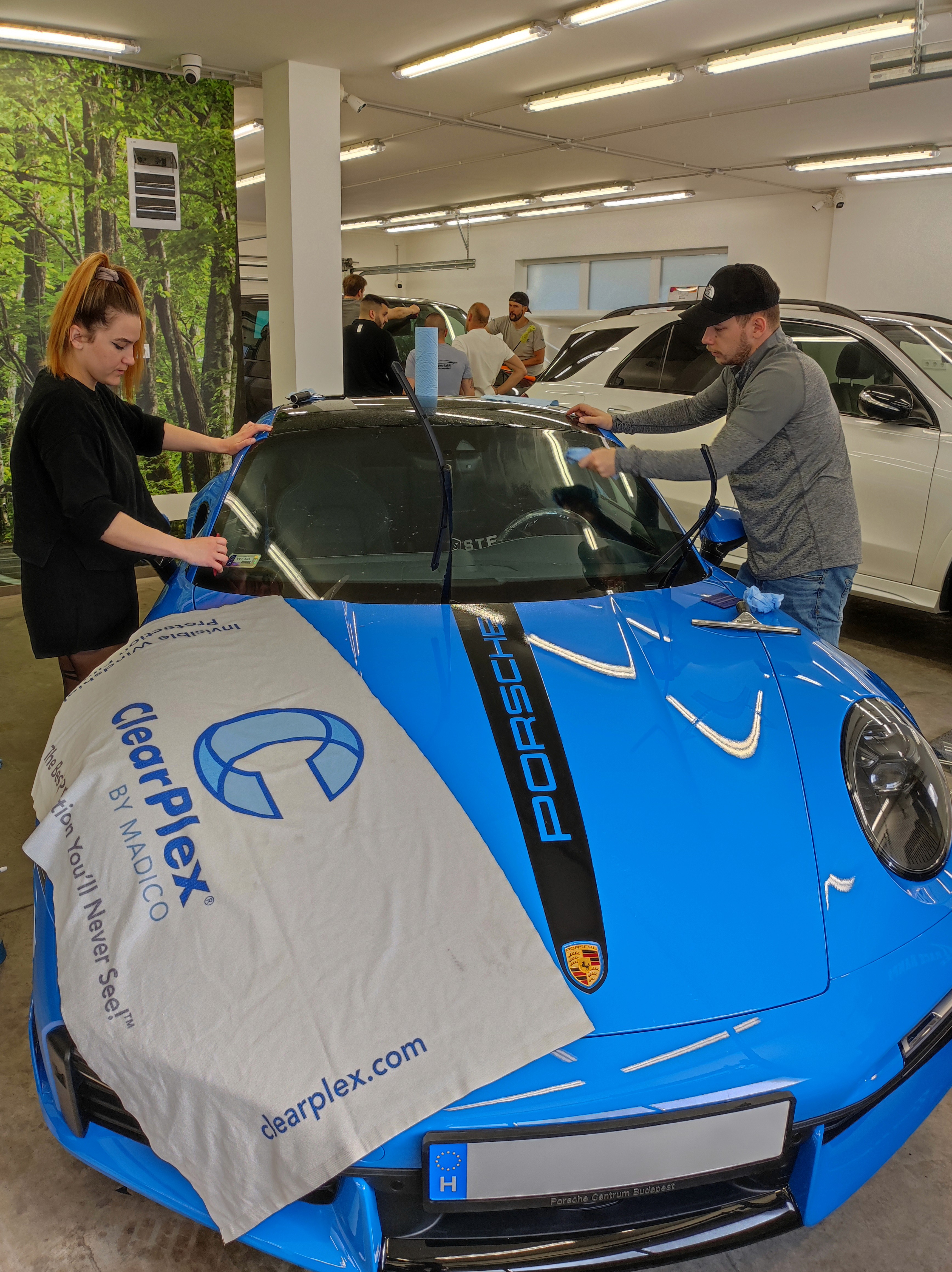 A Nortek szakemberei prémium ClearPlex szélvédővédő fóliát telepítenek egy kék Porsche 911 sportautó szélvédőjére a kőfelverődések és repedések megelőzése érdekében.