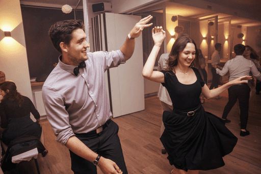Ein junger Mann im Anzug und Fliege und eine junge Frau im schwarzen Kleid tanzen in einem belechteten Raum mit anderen Leuten