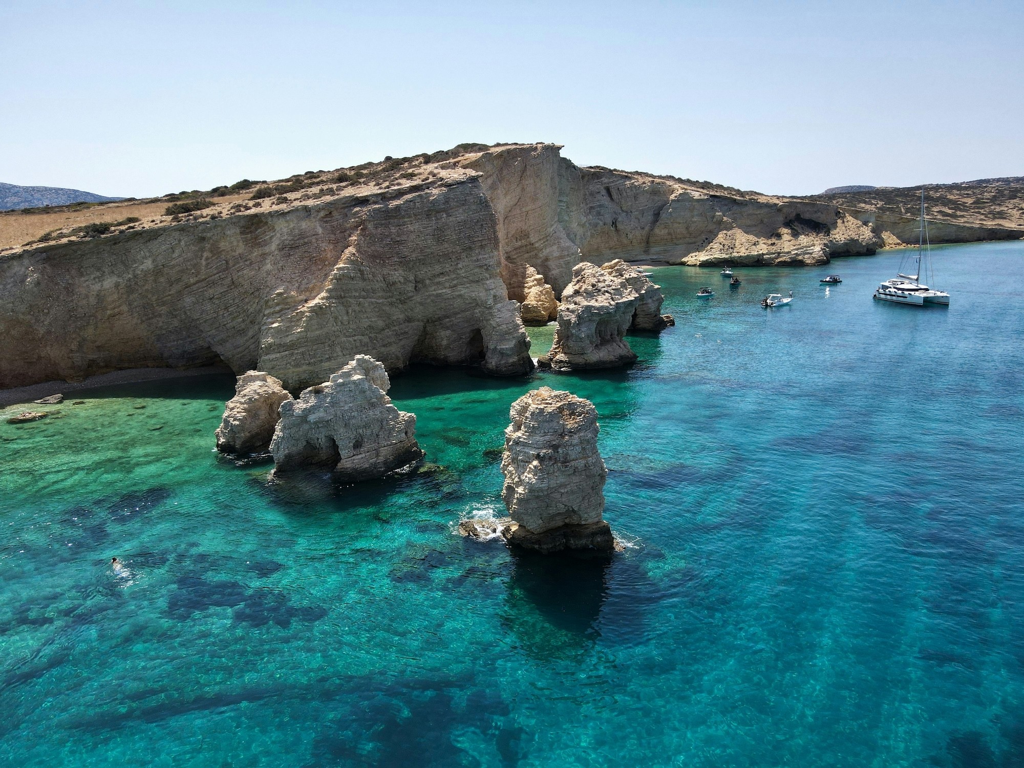Aerial view of dramatic rock formations rising from turquoise waters in a secluded Cycladic cove with boats anchored nearby