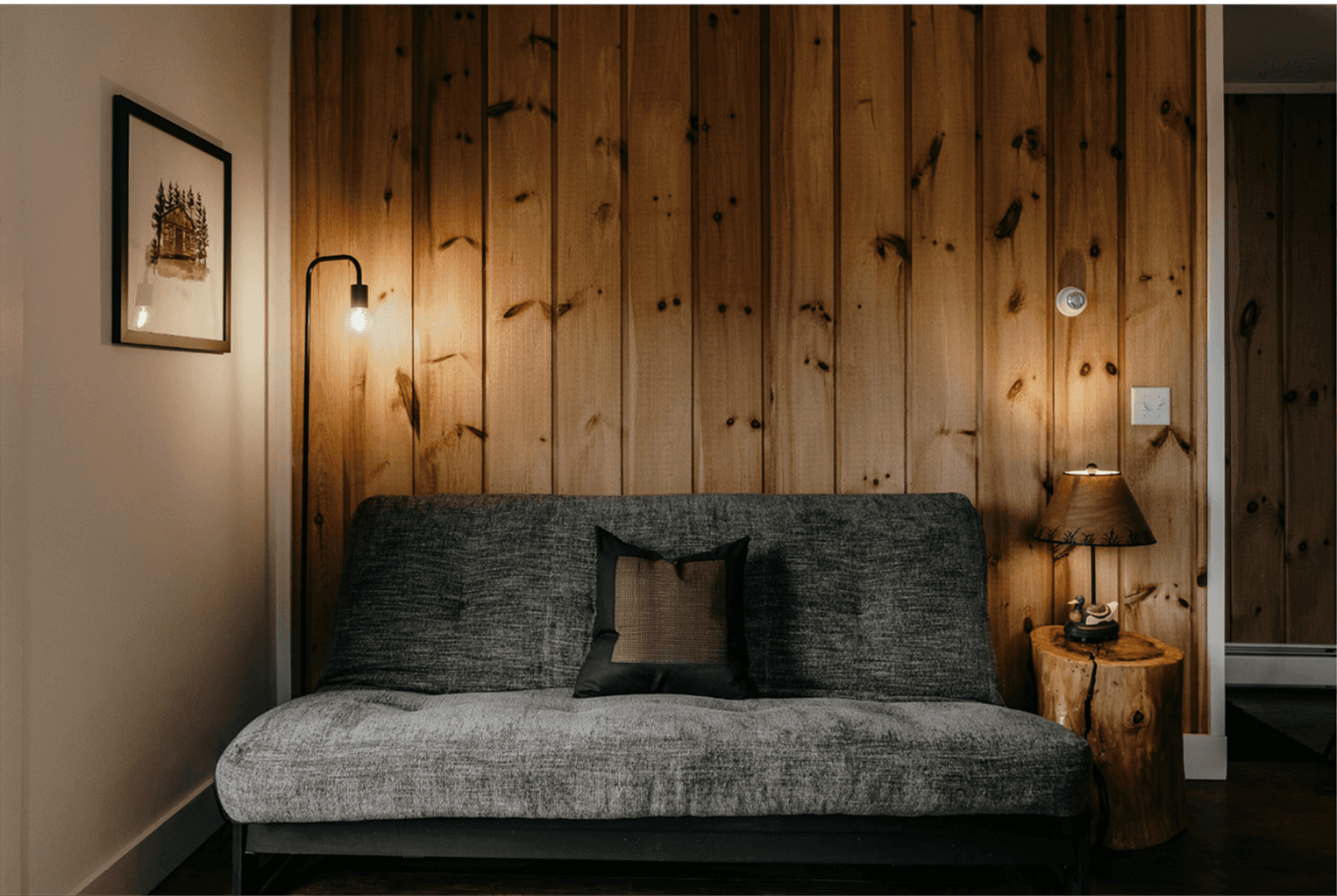 Interior of a Strawtown modular home showing a warm timber-clad wall, pendant bulb lamp, grey sofa, and framed cabin artwork