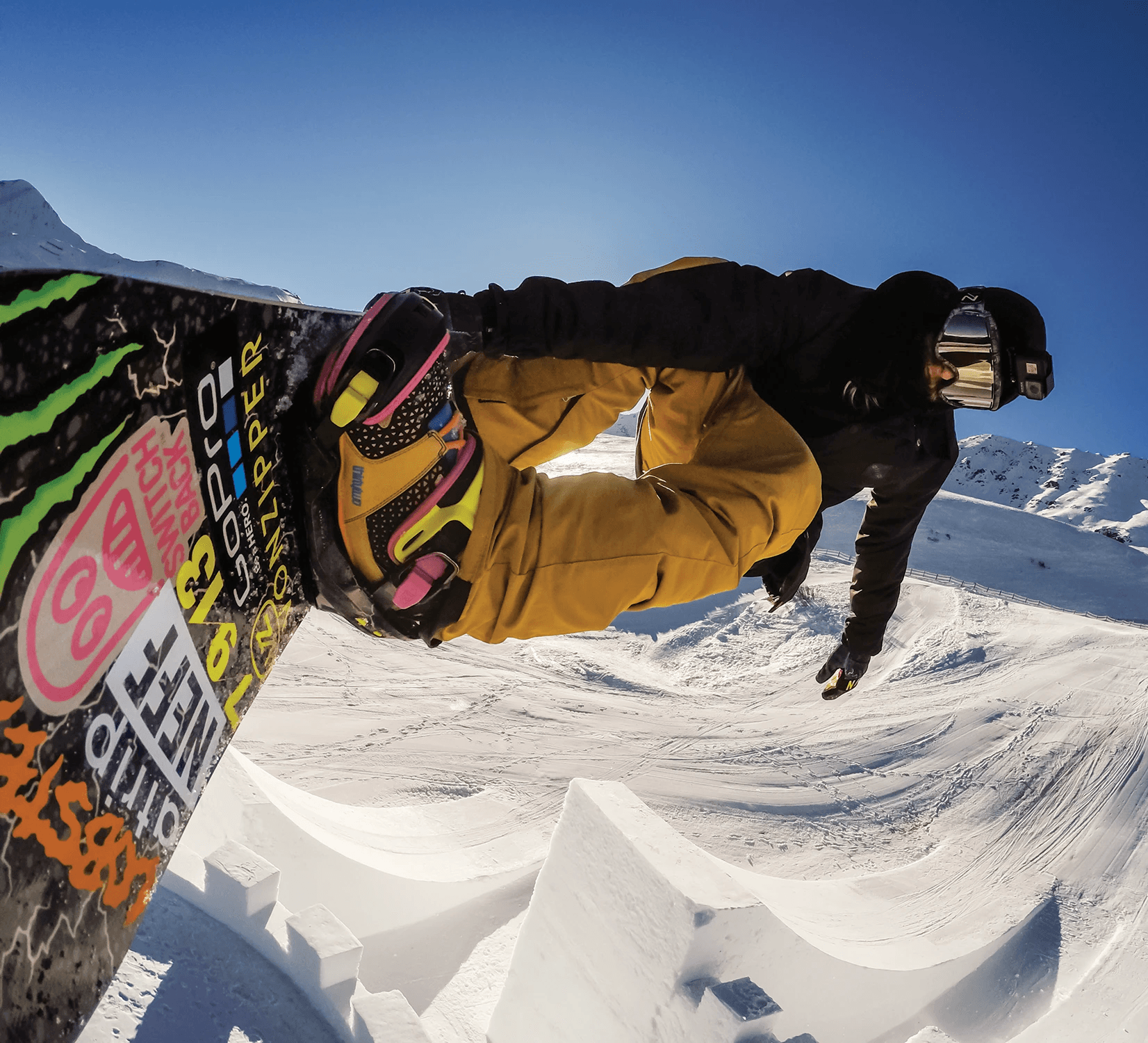 A GoPro camera mounted to the well-stickered board of professional snowboarder Halldor Helgason as he flies over a massive kicker built on a snow-covered Watles Ski Area in the Italian Alps. Halldor is wearing colorful boots and bindings, with tan pants, a black jacket, gloves, and beanie, with reflective snowboarding goggles and another GoPro on his head.