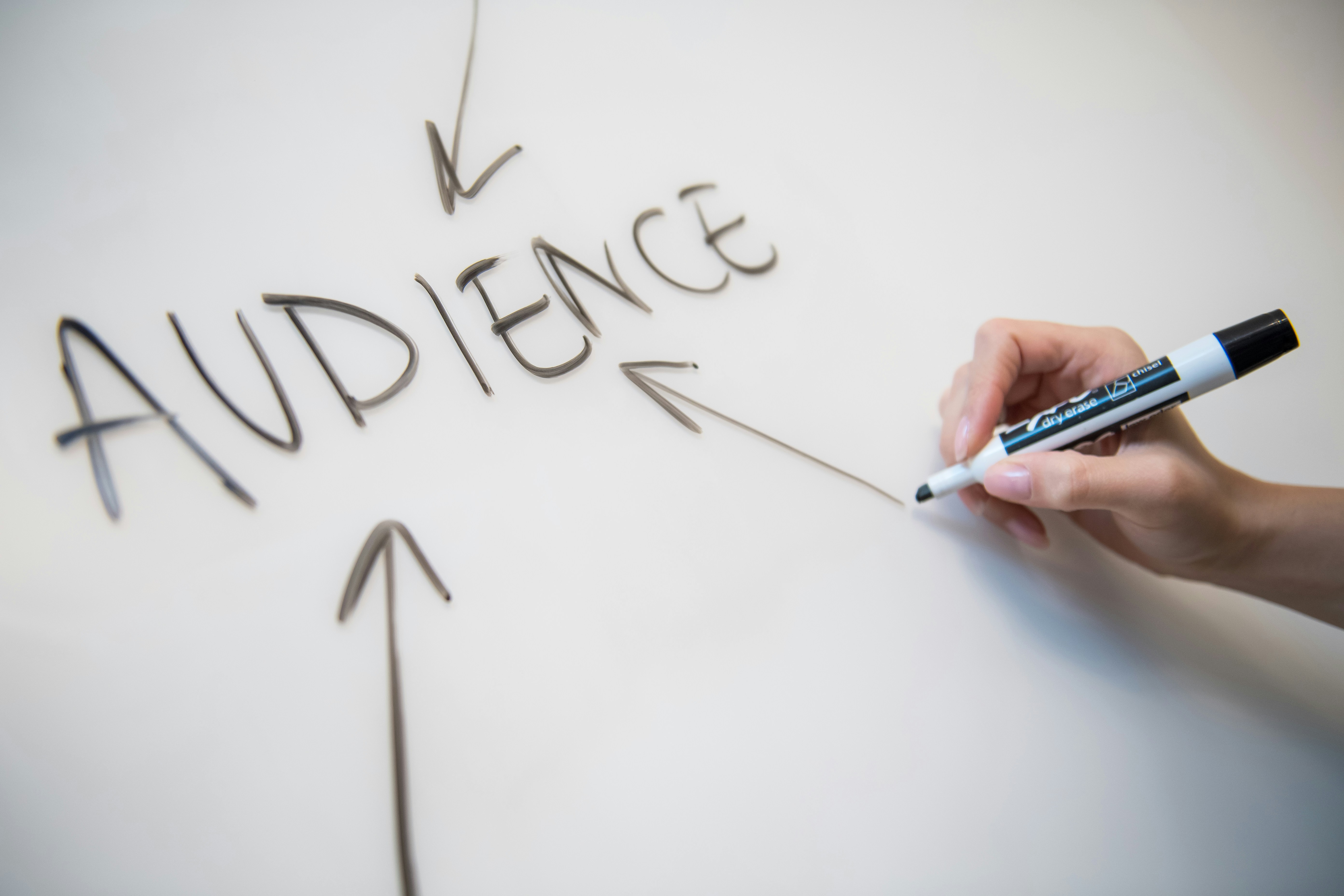 A hand holding a marker writing "AUDIENCE" with arrows pointing upward on a white background.