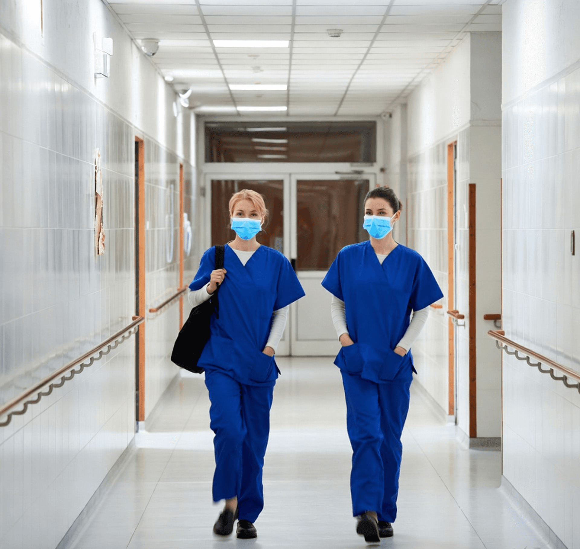Two masked nurses in blue scrubs walk down a hospital corridor.