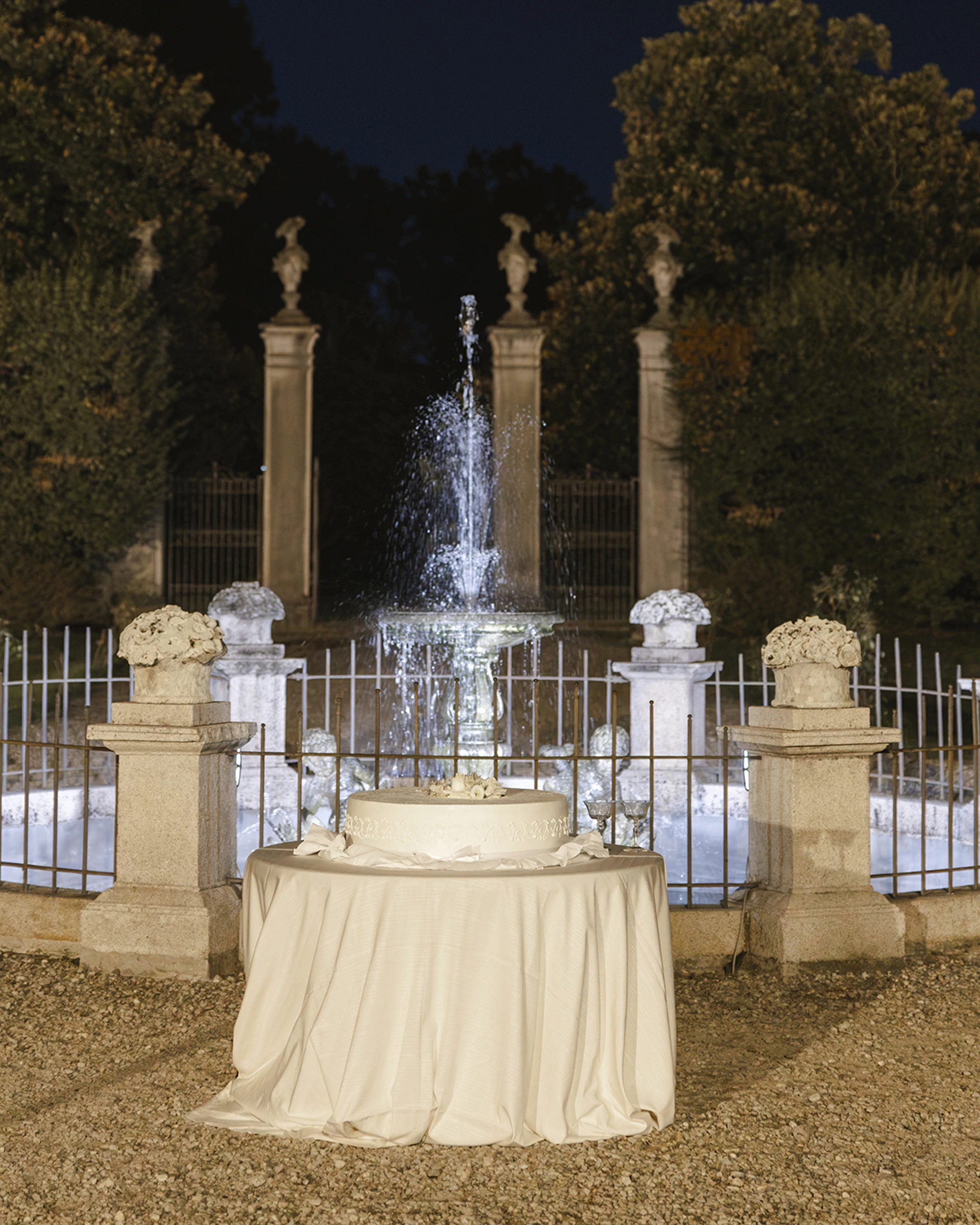 Torta nuziale su tavolo con tovaglia color crema davanti a una fontana illuminata nel giardino, di sera