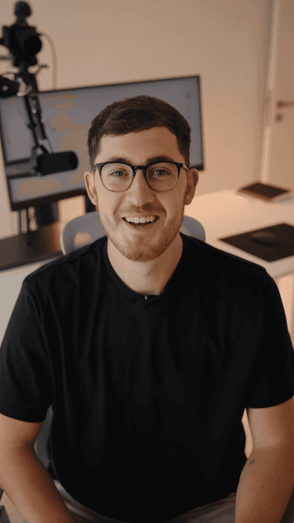 A person wearing glasses is smiling in a modern home office setup with a large monitor, camera, and microphone in the background.