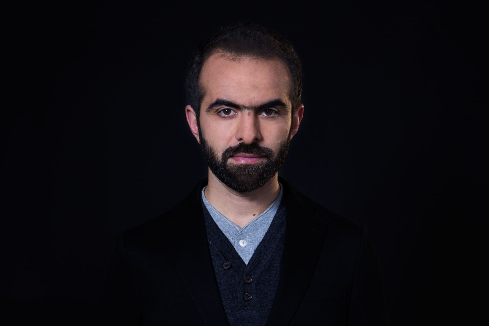 A man with short dark hair and a dark beard is positioned against a black background. He wears a dark blazer over a gray v-neck sweater and a light gray button-down shirt. His expression is serious, and his gaze is directed straight at the camera.