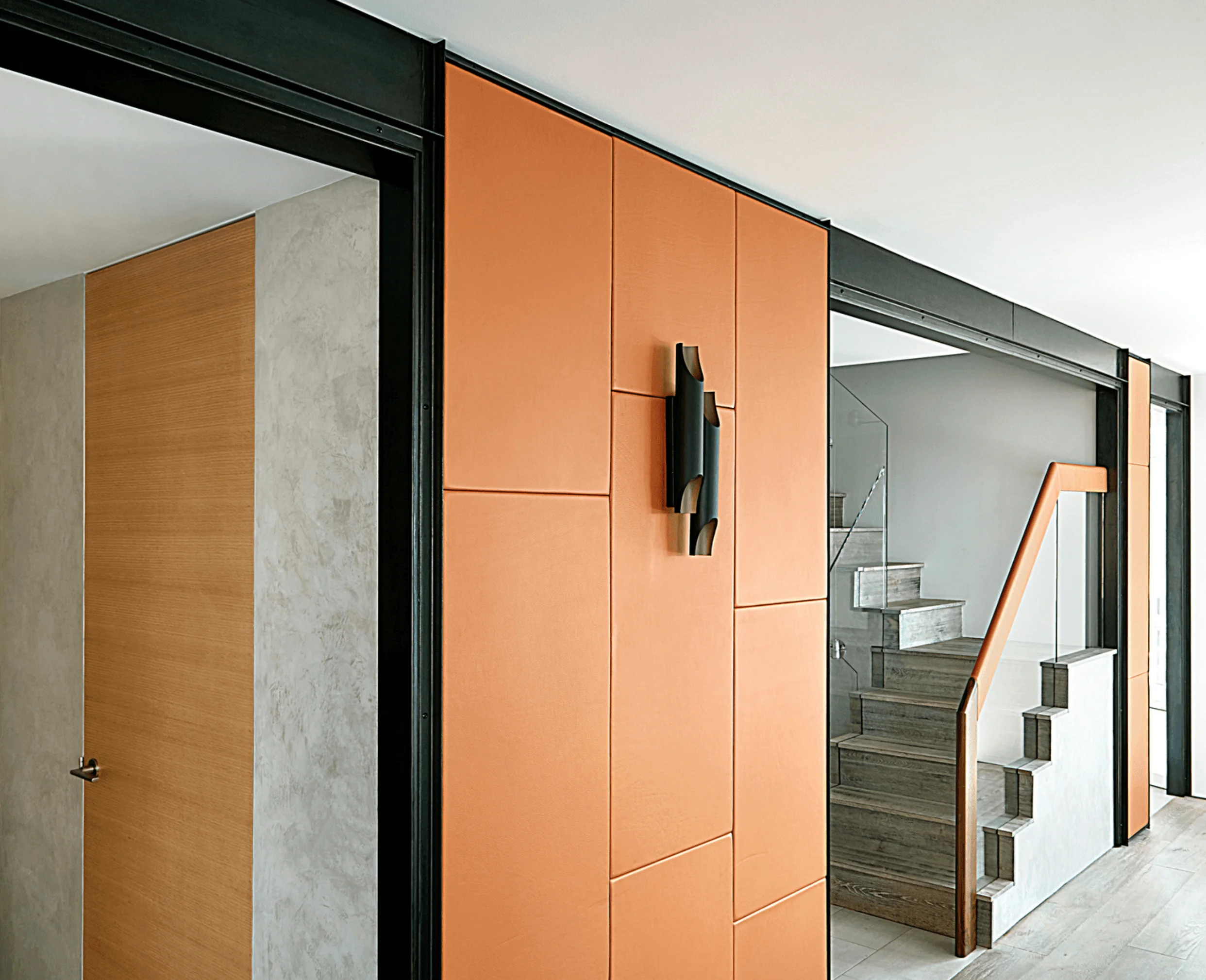 A hallway with a wood door, an orange leather panel wall with a sculptural black sconce, and a glimpse of the staircase beyond