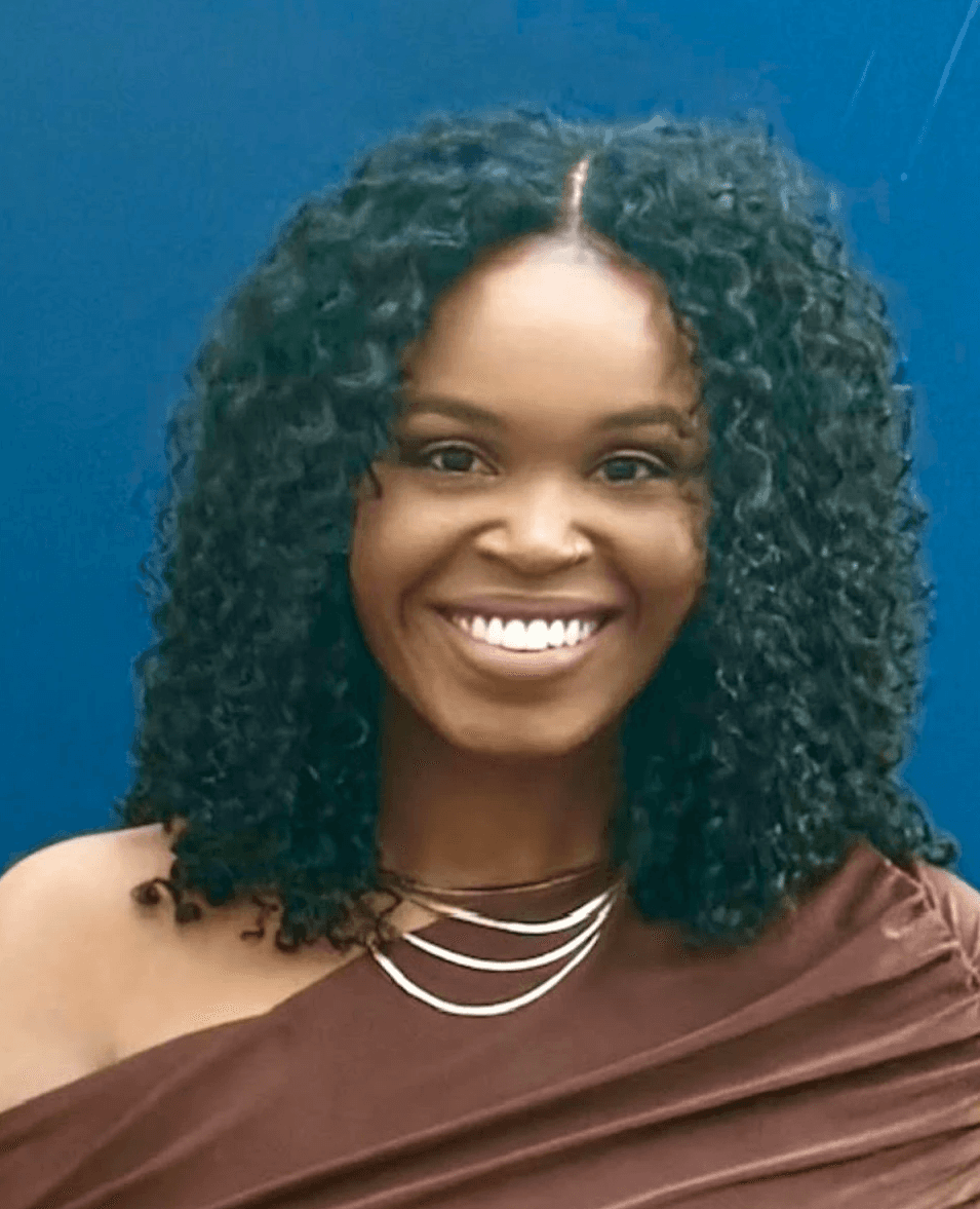 Smiling person with curly hair, wearing a black shirt, against a warm, dark background.