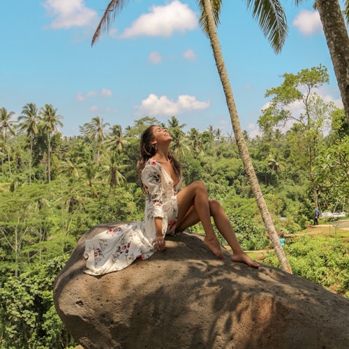 A woman in a floral dress sits on a large rock, surrounded by lush greenery and tall palm trees, under a partly cloudy sky.