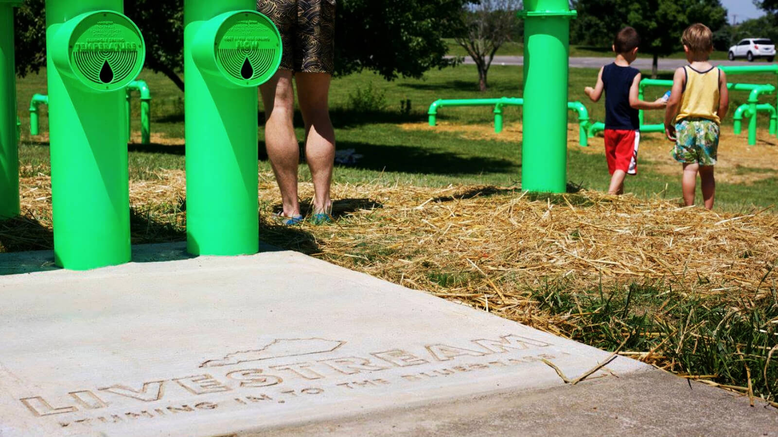 A large interactive sound sculpture weaves in and out of the grass in Jacobson Park. 'LIVESTREAM' is engraved in the concrete leading up to three pipes. Two small children walk toward the pipes. Their father stands back and watches them play.
