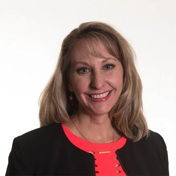 Professional woman with long hair smiling, wearing a black blazer against a dark background. Dr D'Anne Rudden