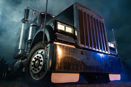 A large, polished semi-truck with headlights on, illuminated against a misty, dark background, showcasing its chrome grille and robust front end.