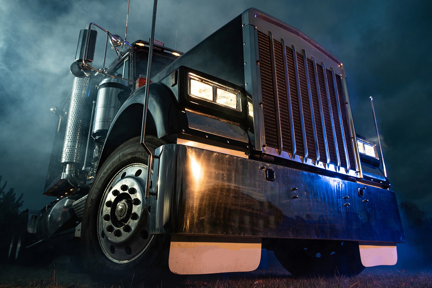 A large, polished semi-truck with headlights on, illuminated against a misty, dark background, showcasing its chrome grille and robust front end.