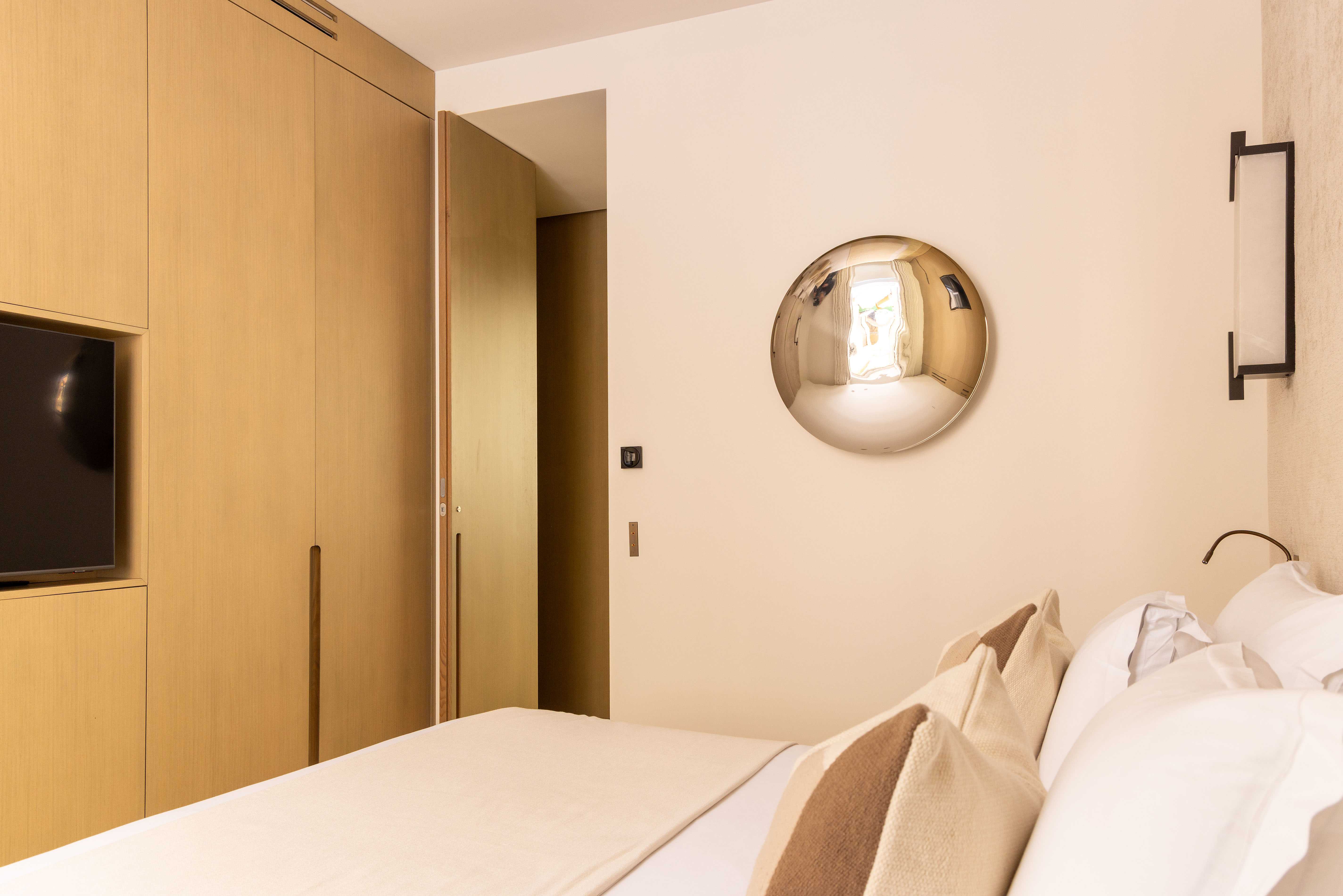 Chambre avec rangements en bois clair, télévision, miroir convexe doré et lit aux tons sable