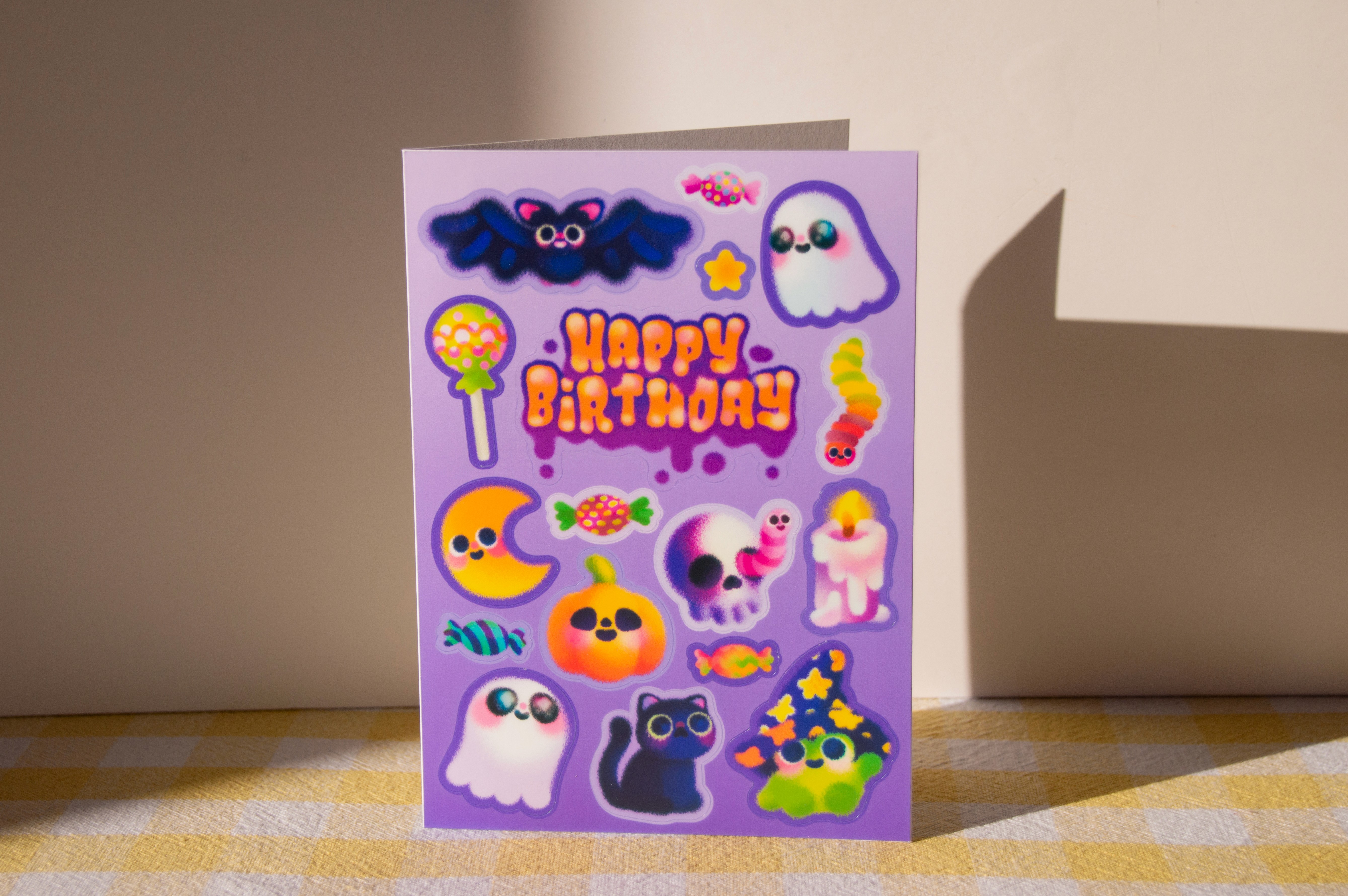 A purple birthday card that is also a sticker sheet, fesaturing halloween themed designs. There are ghosts, a wizard frog, a jack-o-lantern, sweets, and more.