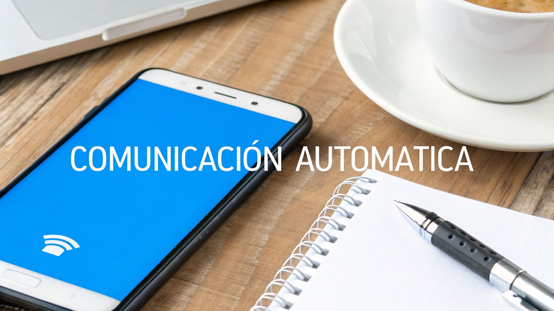 Un smartphone con pantalla azul muestra 'Comunicación Automática' y un icono de señal, en un escritorio de madera.
