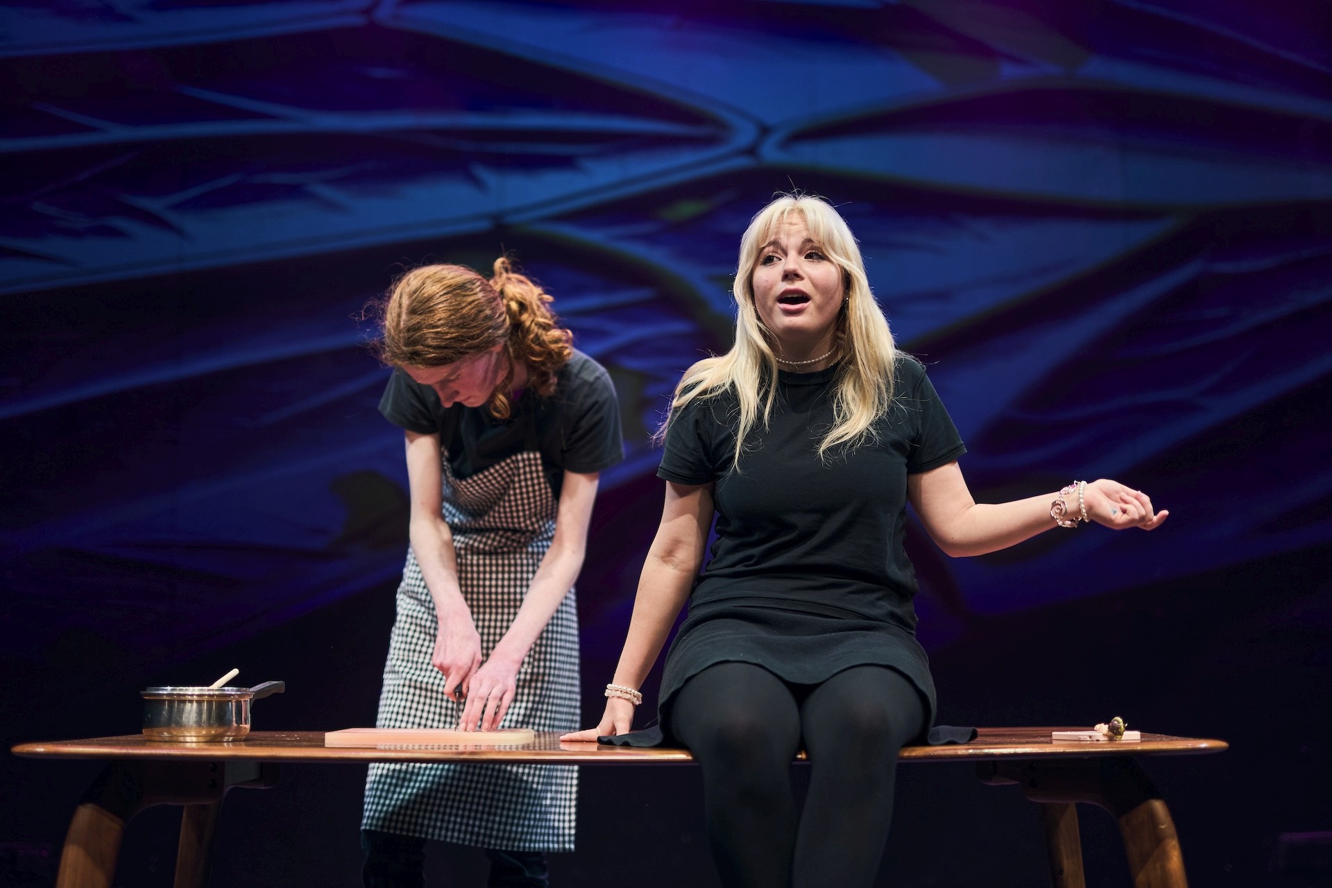 A young performer sits on a wooden bench onstage, mid-monologue, while another performer beside them chops ingredients at a kitchen bench. The scene is theatrical, with dramatic purple lighting and a pot on the table suggesting a cooking setting.