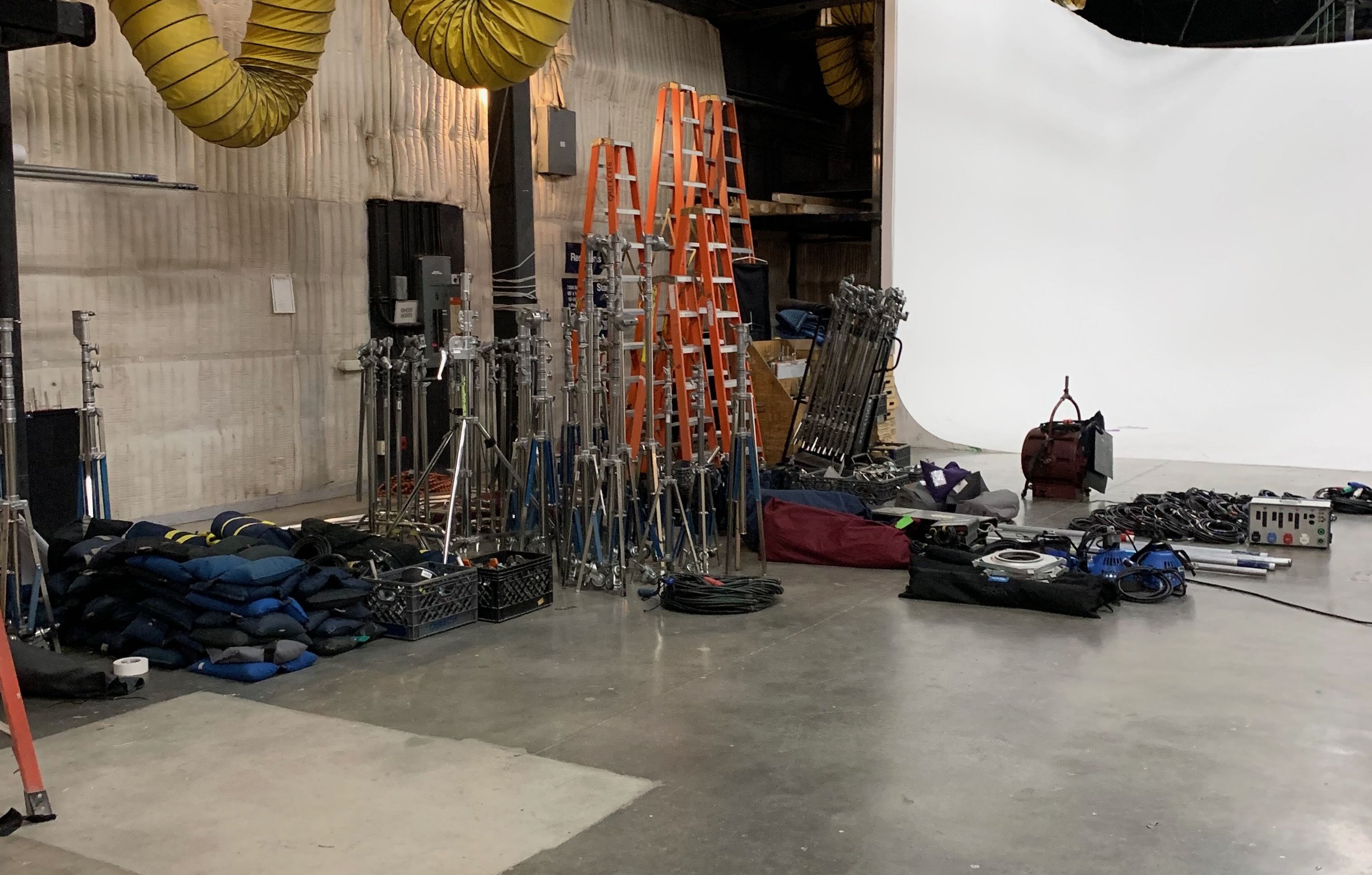 A stage grip package staged on the floor during load-in, including combo stands, orange ladders, sandbags and coiled cable in front of a white cyc wall.