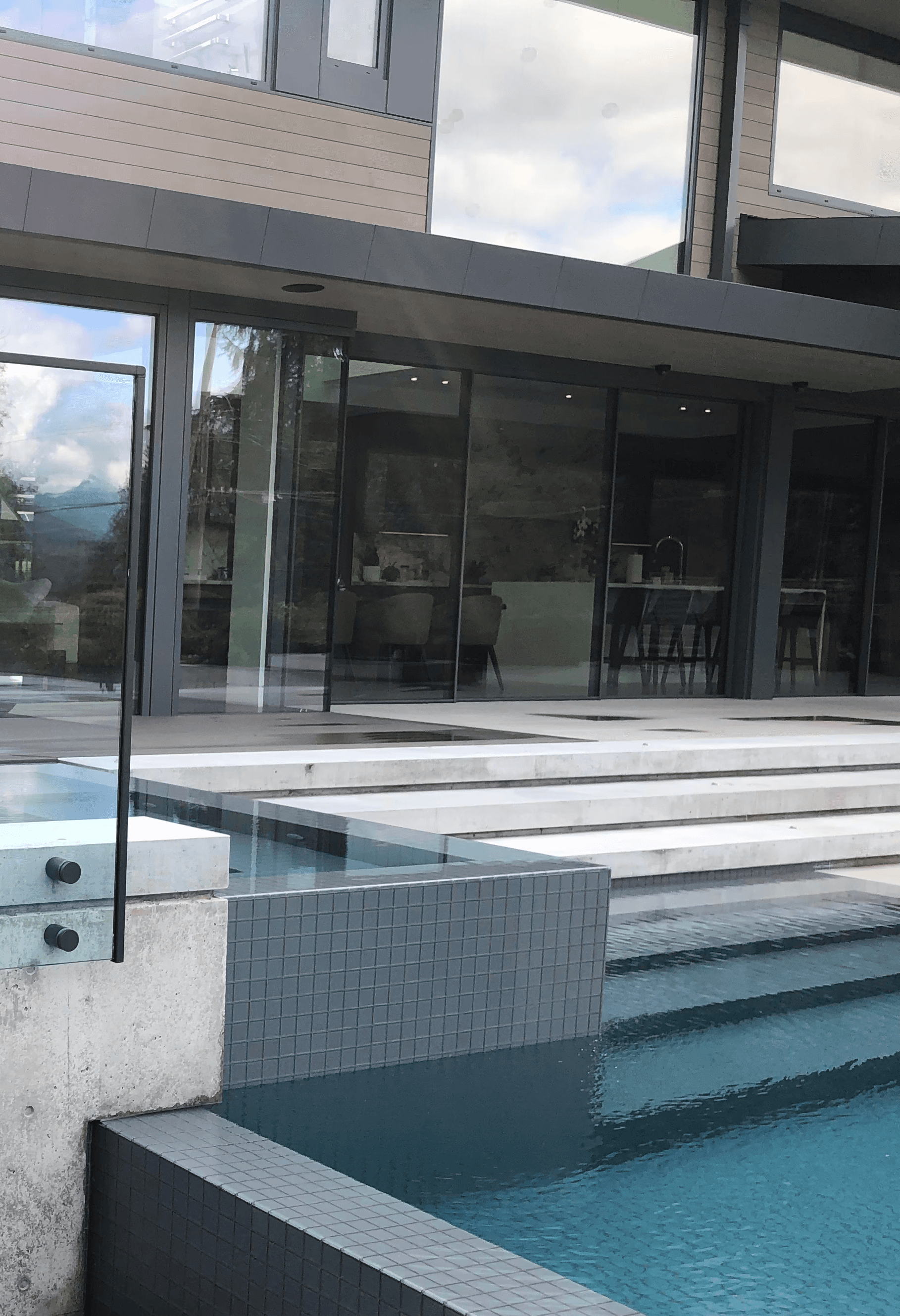 An architectural close-up of a contemporary backyard pool area. A raised concrete hot tub is centered, enclosed by clear glass panels with black spigots. To its right lies a turquoise swimming pool with wide descending steps. A dark timber walkway is visible in the lower left, while the large windows of the modern house reflect the sky and surrounding foliage.