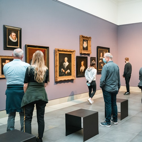 在艺术画廊中，人们观看紫色墙面上的框架肖像，前景中有长椅。