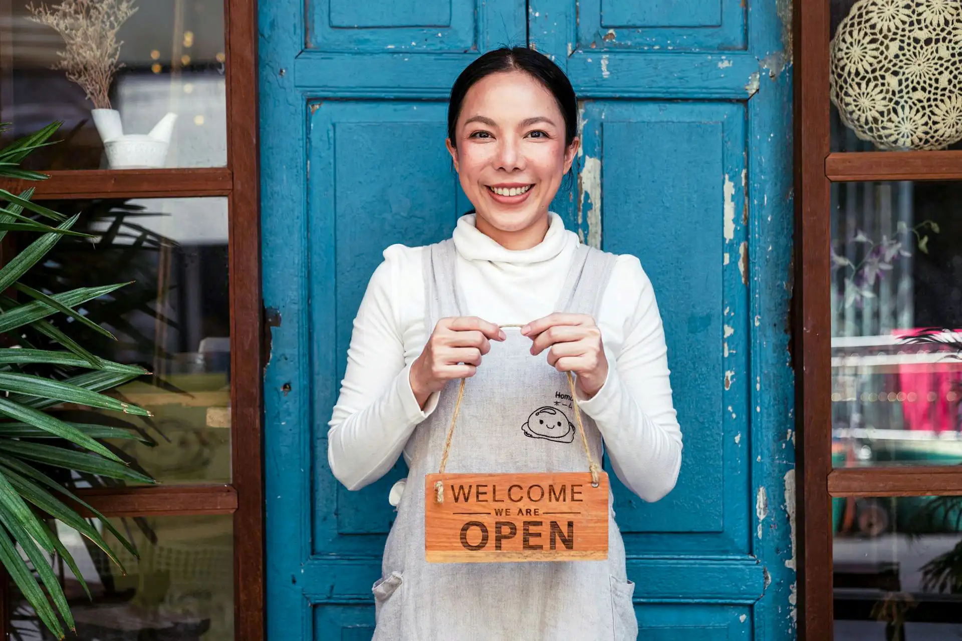 Smiling Asian woman holding "Welcome We Are Open" sign in front of her shop.