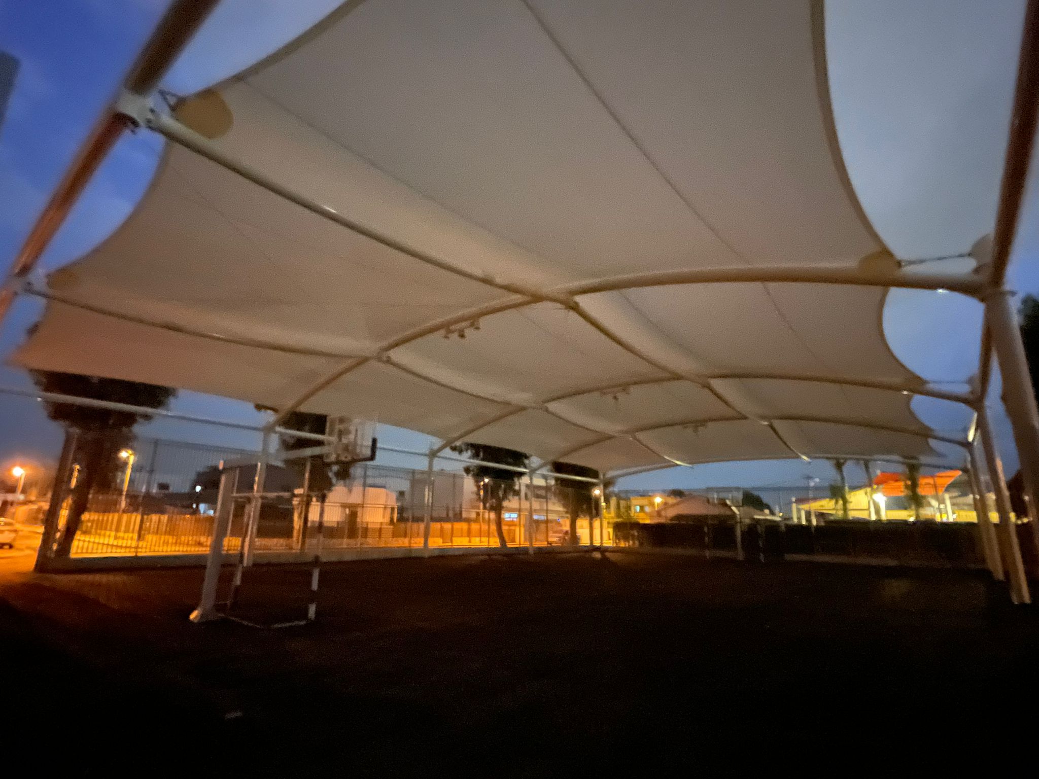 Large beige sunshades over a dark ground at dusk with buildings in the background.
