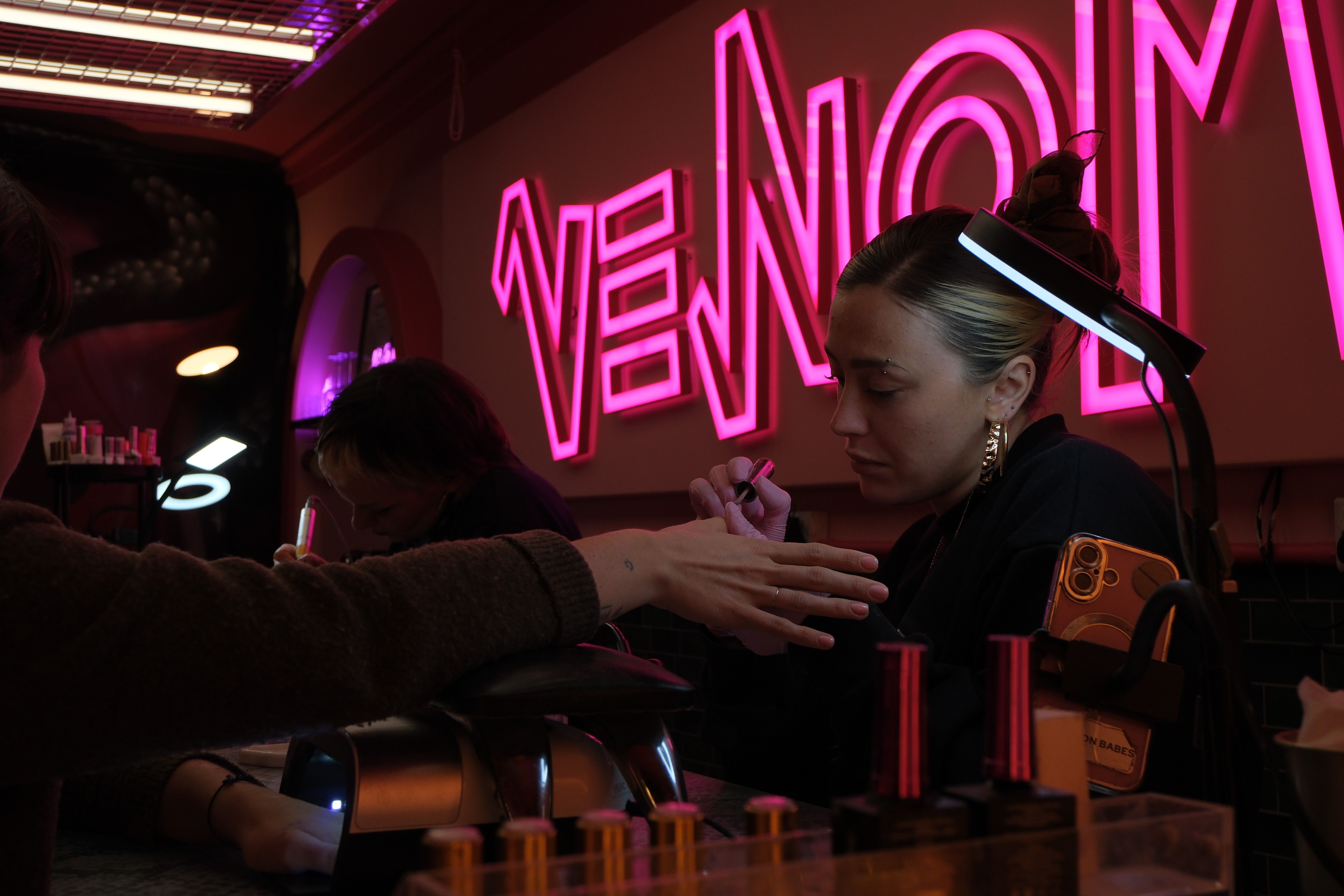 A moody, cinematic shot of a technician carefully applying polish under a bright desk lamp, with the pink "VENOM" neon sign blurred beautifully in the background.