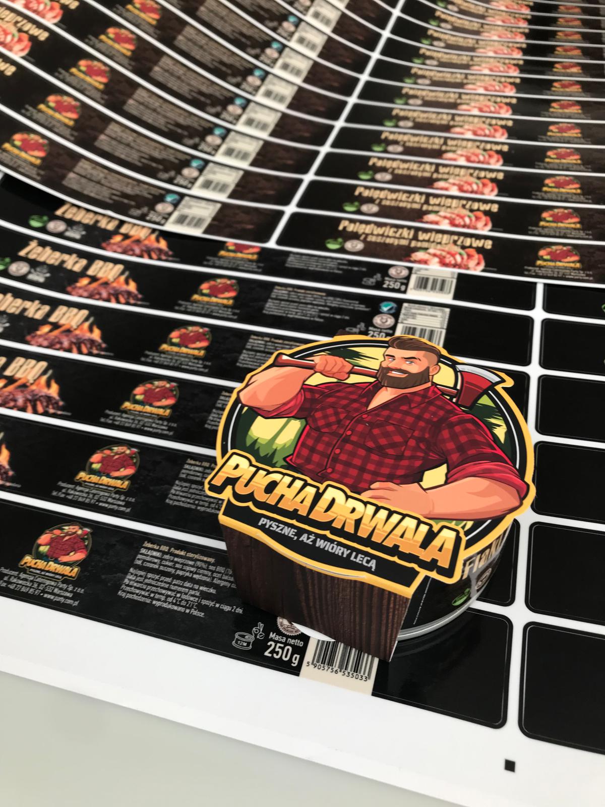 Print Prepress: sheets of colourful packaging labels for "Pucha Drwala" are displayed on a flat surface, featuring vivid graphics of grilled meat and tin cans that highlight the brand and nutritional information.