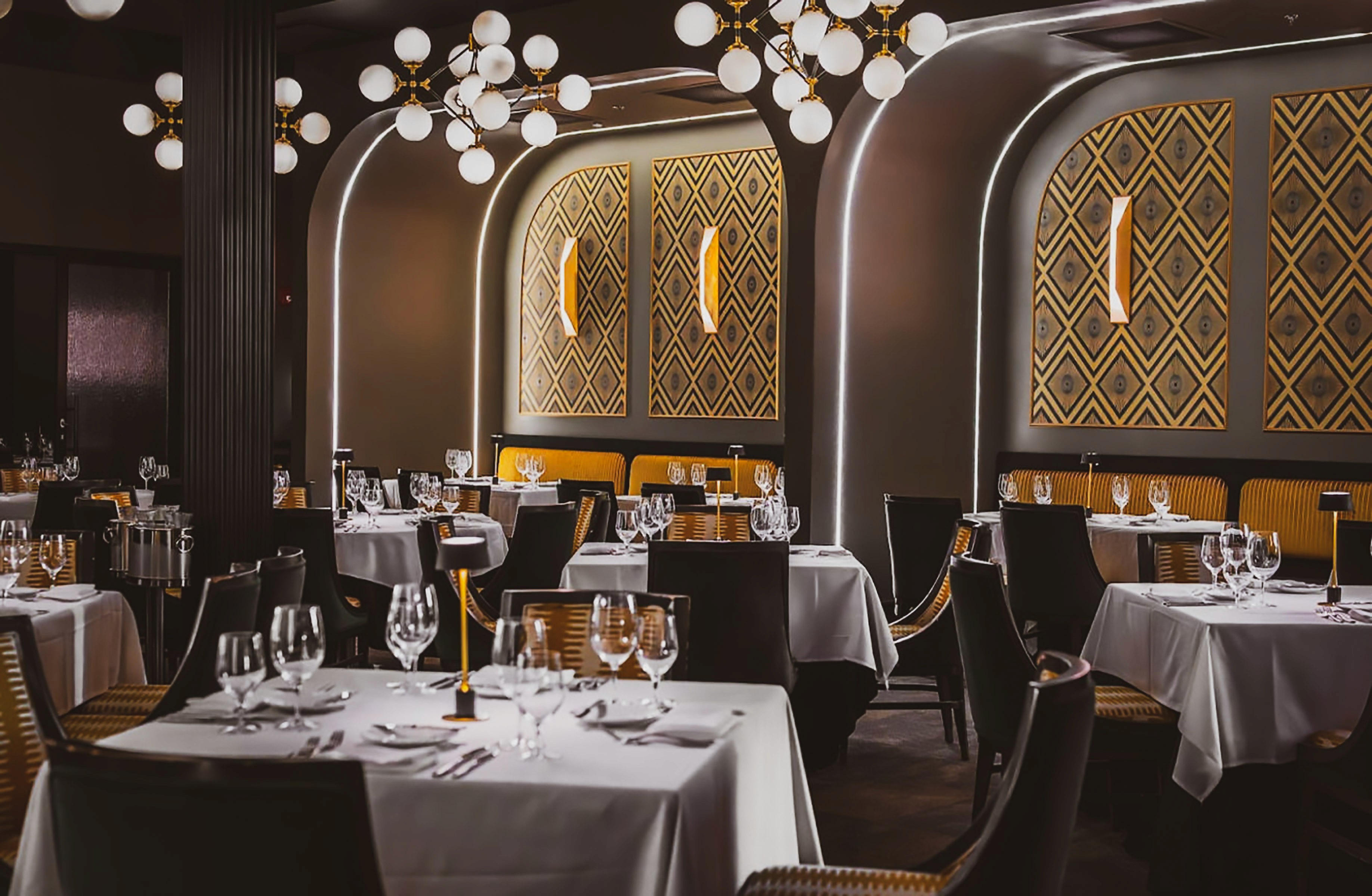 Upscale restaurant interior with elegant tables set for dinner.  Dark walls, gold accents, and modern lighting create a sophisticated ambiance.