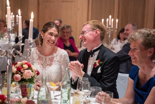 Brautpaar lacht gemeinsam mit Gästen am Tisch während Hochzeitsfeier