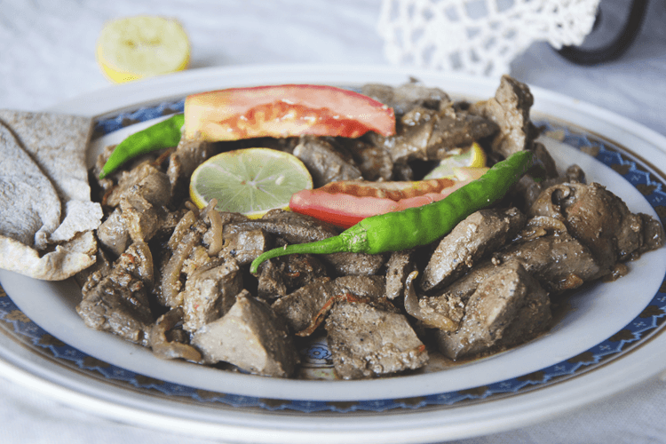 A white plate with a blue and gold rim holds sliced meat, likely beef, garnished with tomato slices, a lime wedge, and a green chili pepper. A piece of light-colored flatbread is visible to the left of the plate, and a lemon wedge rests on the table beside it. The plate is positioned on a white tablecloth, with a white doily or lace item partially visible in the background.