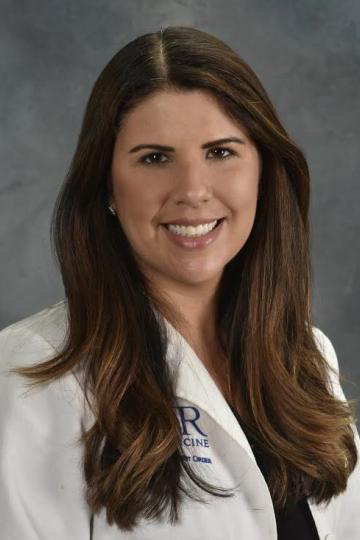 Portrait of a smiling woman with long brown hair, wearing a white lab coat, likely a doctor or medical professional.