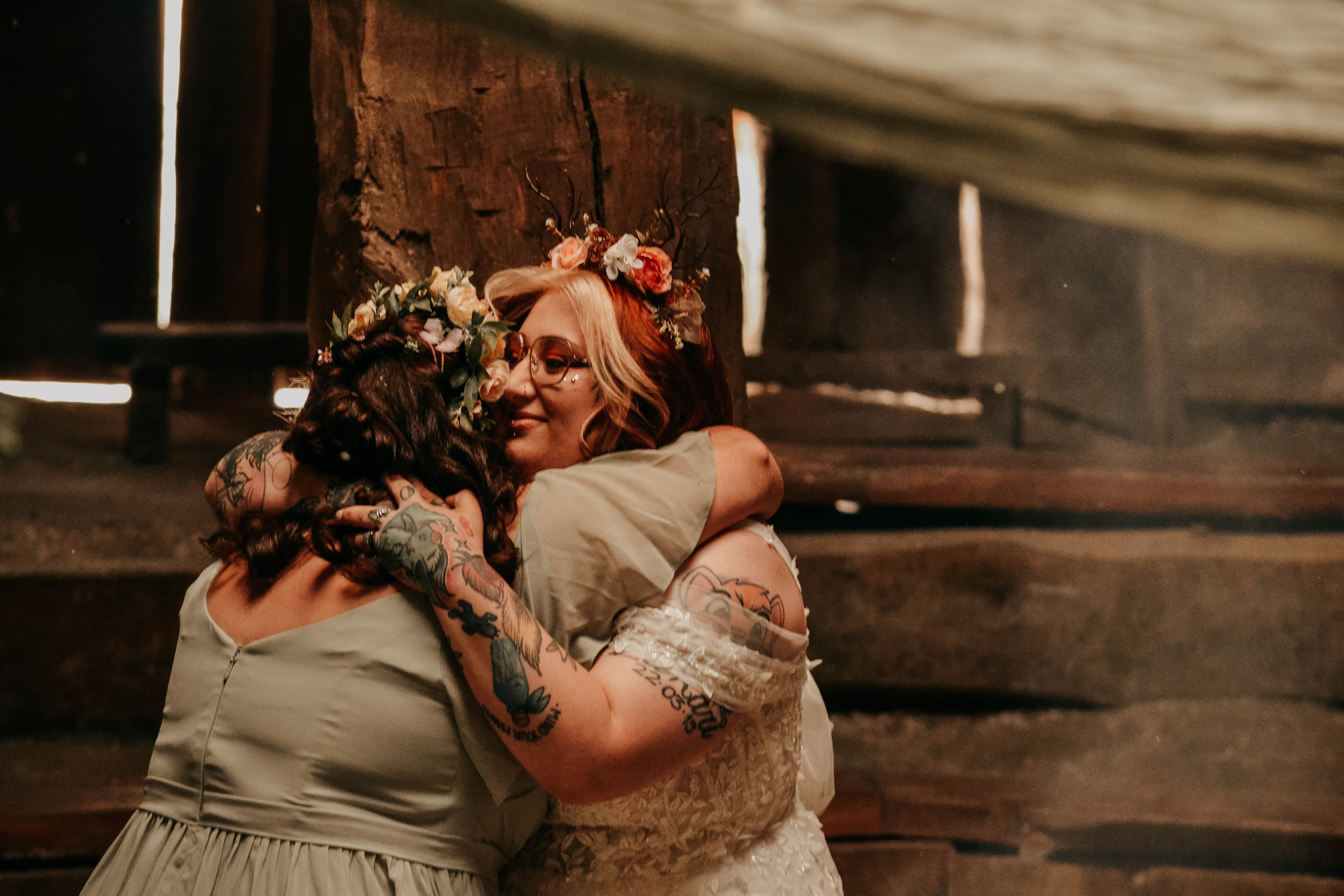 A bride with red hair, a floral twig crown, and tattooed arms shares a warm hug with another woman in a rustic, wooden setting. The bride is smiling with her eyes closed in a moment of affection.