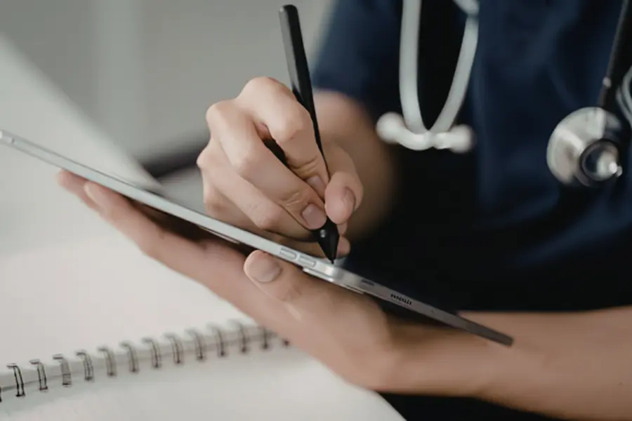 Medical coder AI simplifying charting as a healthcare worker in navy scrubs writes on a tablet with a stylus.