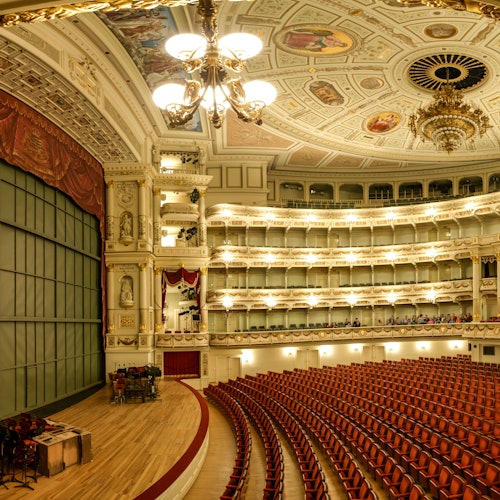 Ylellinen teatteri, jossa on koristeellinen katto, kattokruunu, parvekkeet, punaiset tuolit ja näyttämö, jossa on punainen esirippu.