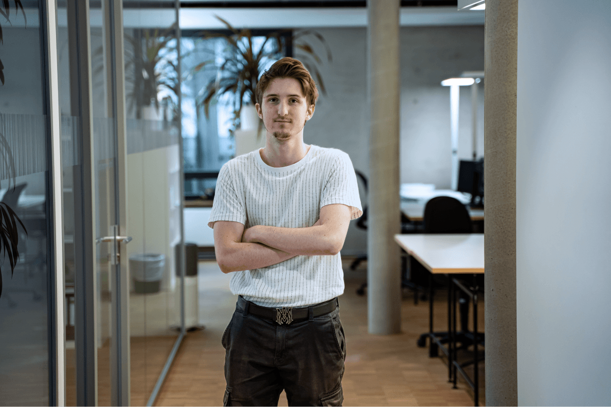 Junger Mann mit verschränkten Armen steht in einem modernen Büroflur der kba Architekten und Ingenieure GmbH; im Hintergrund sind Glaswände, Pflanzen und Arbeitsplätze zu sehen.