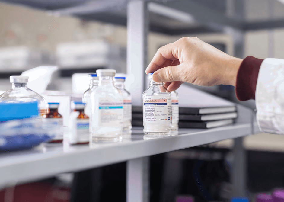 A close-up of a scientist's hand reaching for a labeled pharmaceutical vial from a laboratory shelf.