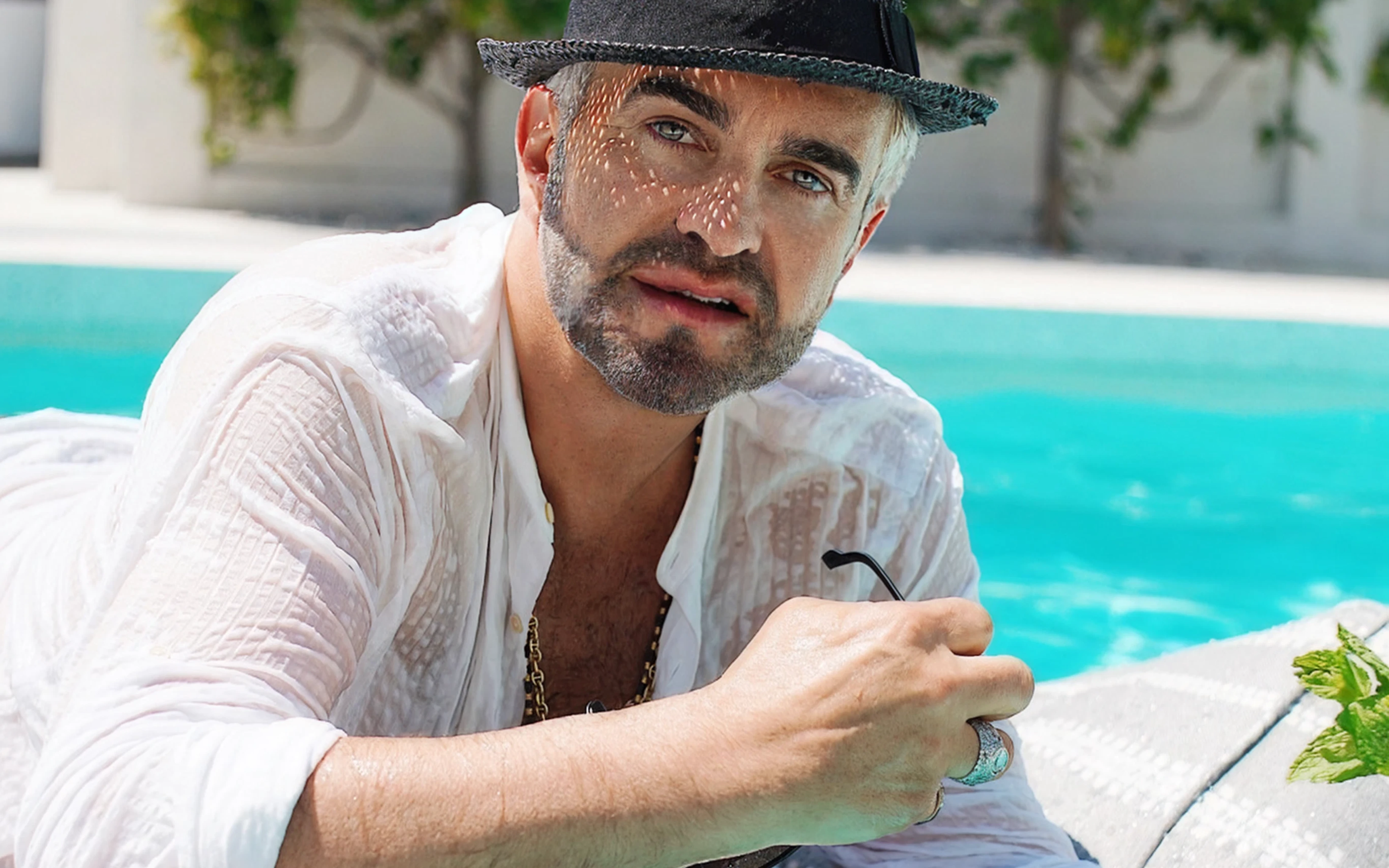 Stylish man in a black hat and white shirt relaxing poolside with Oliver James Lilos — capturing the spirit of slow luxury and effortless confidence.